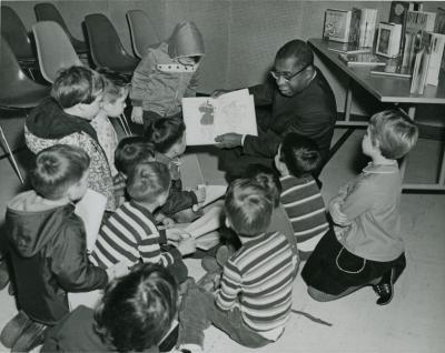 Black and white photograph showing Spencer G. Shaw in formal attire with glasses reading a picture book to a group of young children gathered in a library setting.